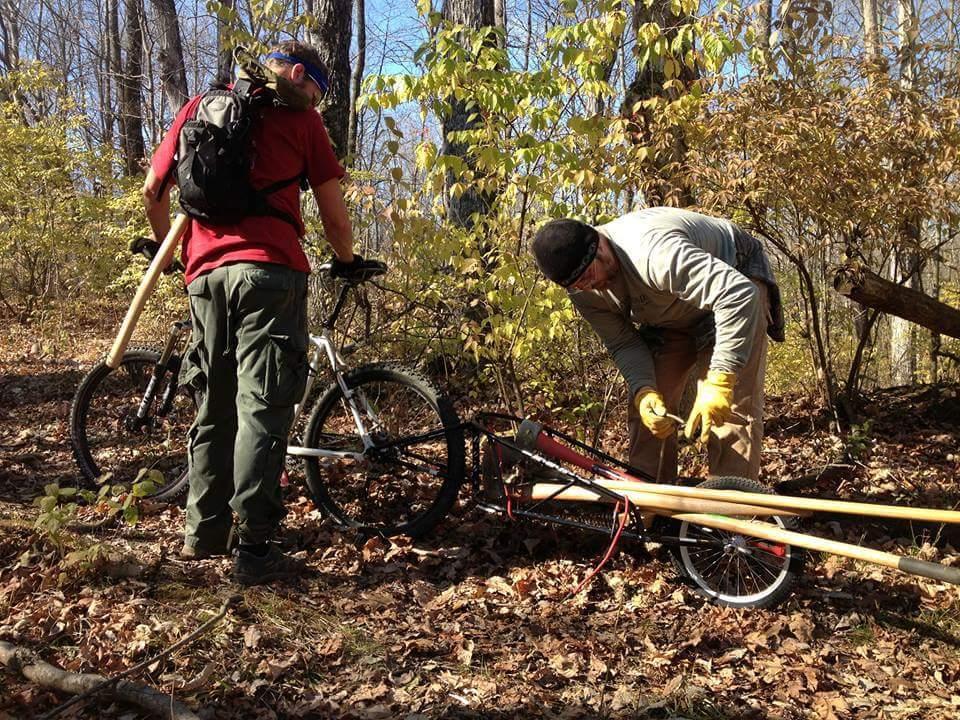 Two individuals working in a wooded area, with one person repairing a bicycle and the other holding a tool. The setting features autumn foliage, fallen leaves on the ground, and two bicycles nearby. The person repairing the bike is wearing a light-colored long-sleeve shirt and gloves, while the other is wearing a red shirt and cargo pants. Hueston Woods State Park mountain bike trail.