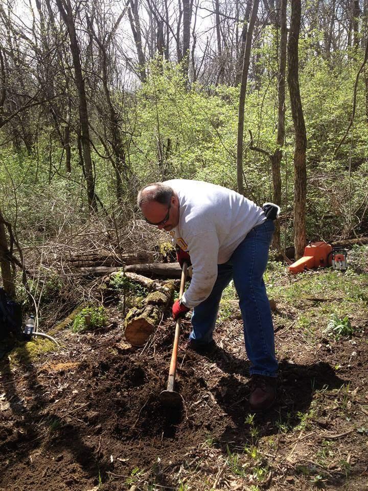 A person is digging in the ground in a wooded area, wearing a light-colored sweatshirt and blue jeans, using a long-handled shovel. Surrounding the individual are trees with budding leaves, and there are fallen logs and greenery in the background. Hueston Woods State Park mountain bike trail.
