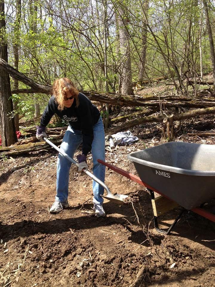 A person using a shovel to dig in the soil in a wooded area, with a wheelbarrow nearby. Sunlight filters through the trees, illuminating the scene. The individual is dressed in a black sweatshirt and jeans, focused on their work. Hueston Woods State Park mountain bike trail.