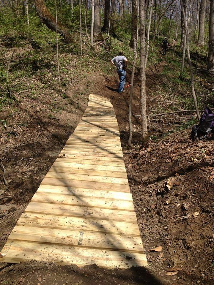 A wooden pathway being constructed in a forested area, with a person working on it. The path is surrounded by trees and green vegetation, indicating a naturistic environment. Hueston Woods State Park mountain bike trail.