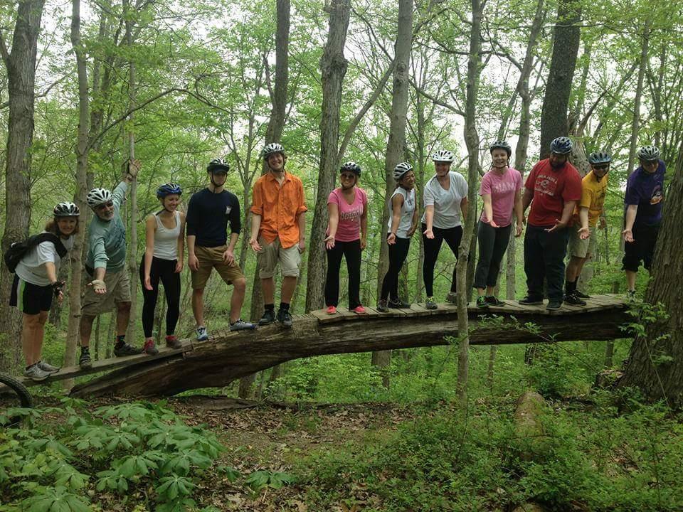 A group of 12 people are standing in a line on a fallen log in a lush green forest. They are wearing helmets and sporting casual outdoor clothing, with some appearing to be posing playfully with their arms outstretched. The scene captures a camaraderie among friends enjoying an adventure in nature. Tall trees and green foliage provide a vibrant backdrop. Hueston Woods State Park mountain bike trail.