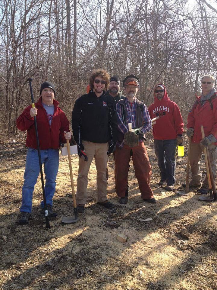 A group of six men standing together in a wooded area, all smiling and holding various tools for outdoor work. They appear to be engaged in a community service or conservation project, with some wearing sunglasses and work gear. The background features bare trees and the ground is covered with sawdust and leaves. Hueston Woods State Park mountain bike trail.