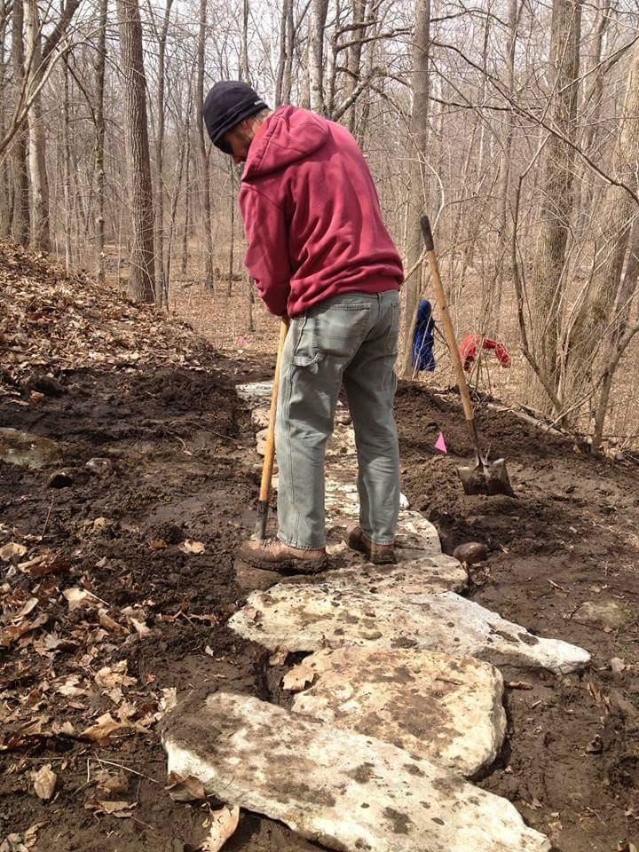 A person in a hoodie and beanie is working on a trail, using a shovel to place large stone slabs in a muddy path surrounded by trees in a forested area. Hueston Woods State Park mountain bike trail.