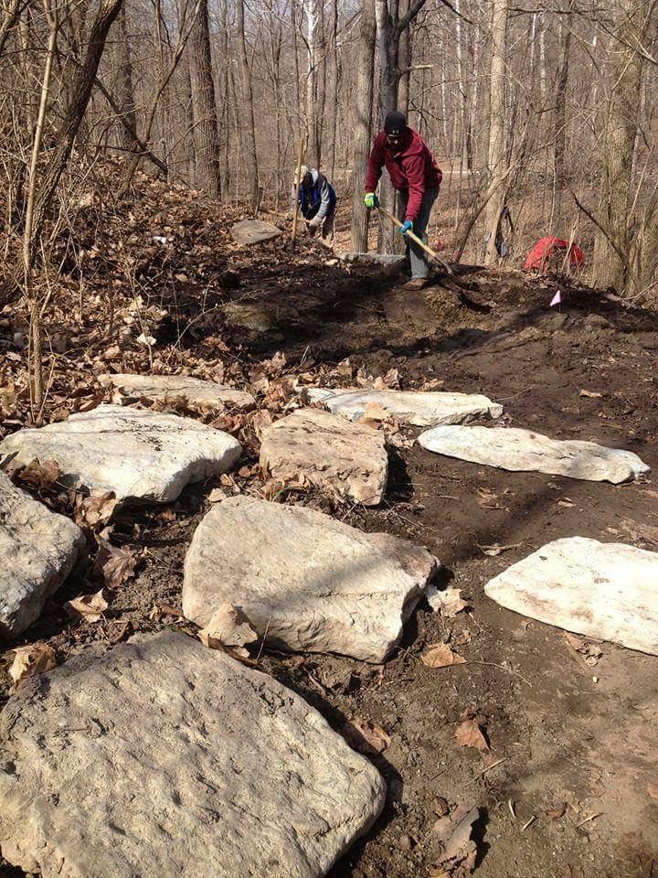 A group of people working on a trail in a wooded area, with large stones laid out on the ground among fallen leaves. One person in the foreground is using a shovel, while another is digging in the background. The scene captures a volunteer effort for trail maintenance or construction. Hueston Woods State Park mountain bike trail.