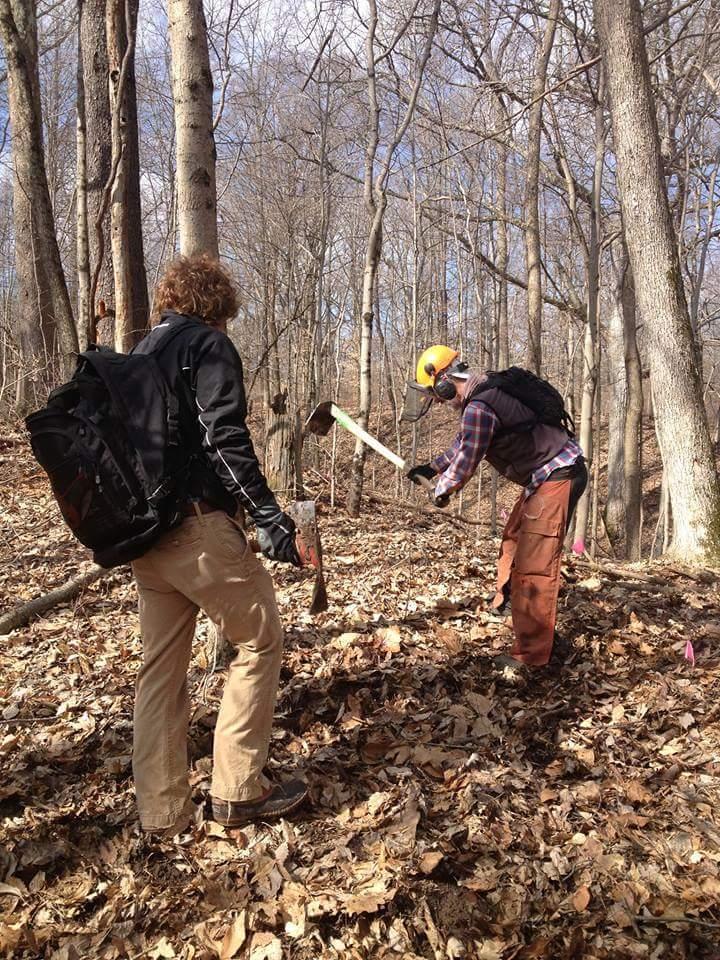 Two individuals engaged in outdoor work in a wooded area. One person is using a large tool while wearing a safety helmet, and the other is observing or assisting. The ground is covered with fallen leaves, and there are trees in the background. Bright sunlight is filtering through the branches. Hueston Woods State Park mountain bike trail.
