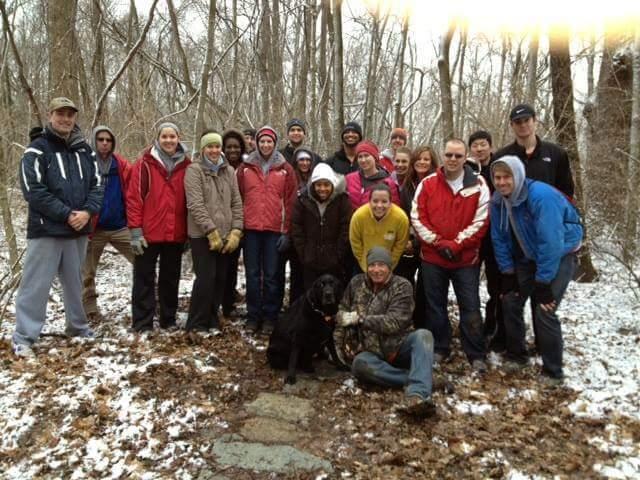 A diverse group of around 25 people is gathered outdoors in a wooded area during winter, with patches of snow on the ground. They are wearing colorful winter jackets, hats, and gloves, indicating cold weather. A black dog is sitting in front of a man who is crouching down. The background features bare trees, creating a serene natural setting. Hueston Woods State Park mountain bike trail.