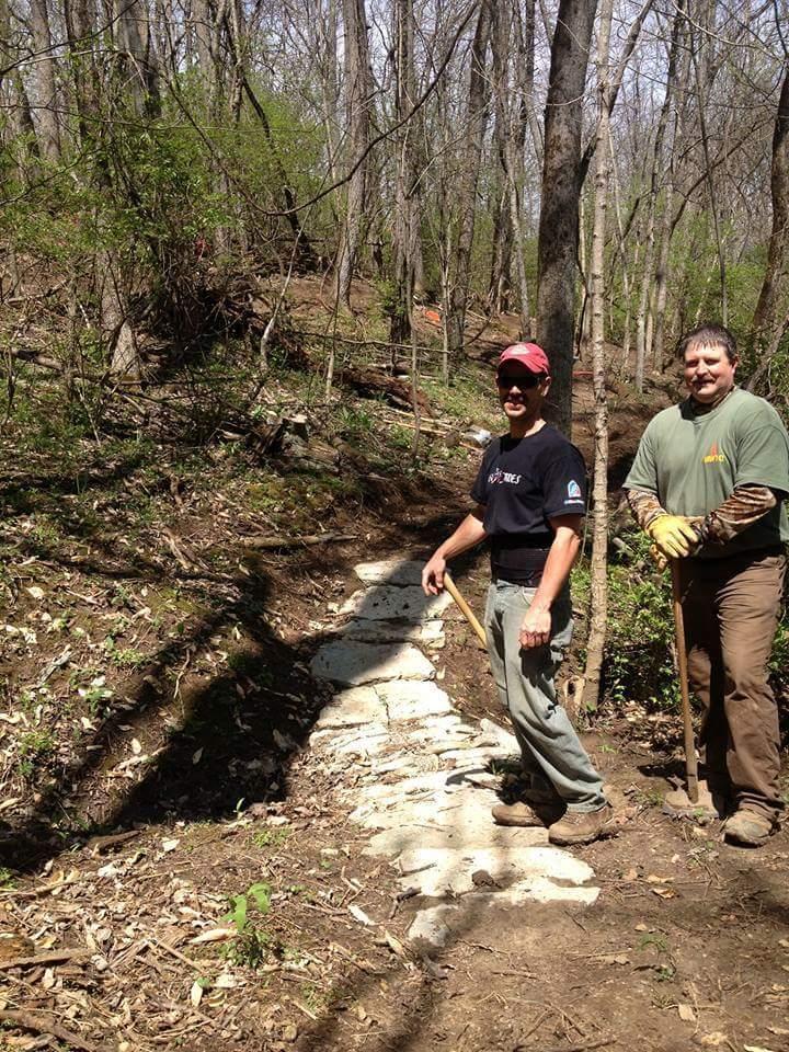 Two men standing on a partly cleared trail in a forested area. One man is holding a tool and wearing a baseball cap, while the other man is wearing gloves and a t-shirt. The background includes trees and underbrush, suggesting an outdoor setting where trail maintenance or construction is taking place. Hueston Woods State Park mountain bike trail.