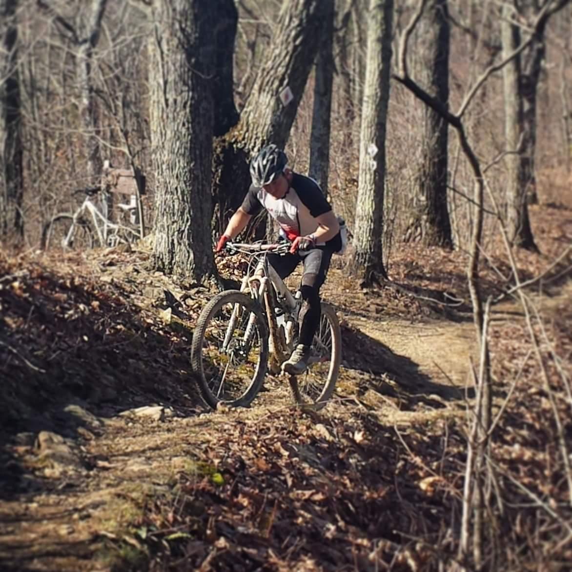 A mountain biker navigating a dirt trail through a wooded area, surrounded by trees and dry leaves. The cyclist is wearing a helmet and is focused on the path ahead, with a second bike visible in the background. Cave-run Lake mountain bike trail.