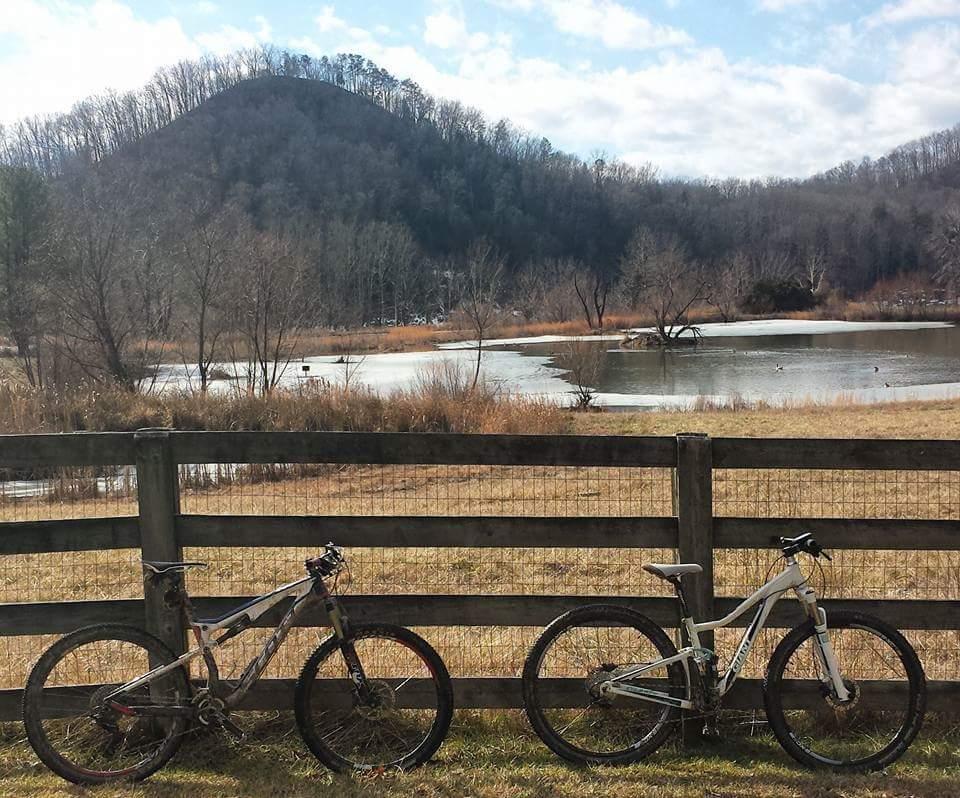 Two mountain bikes are leaning against a wooden fence in a scenic outdoor setting. In the background, there is a calm pond surrounded by tall grasses and a wooded hill. The sky is partly cloudy, and the landscape suggests a tranquil, natural environment with hints of winter still visible. Cave-run Lake mountain bike trail.