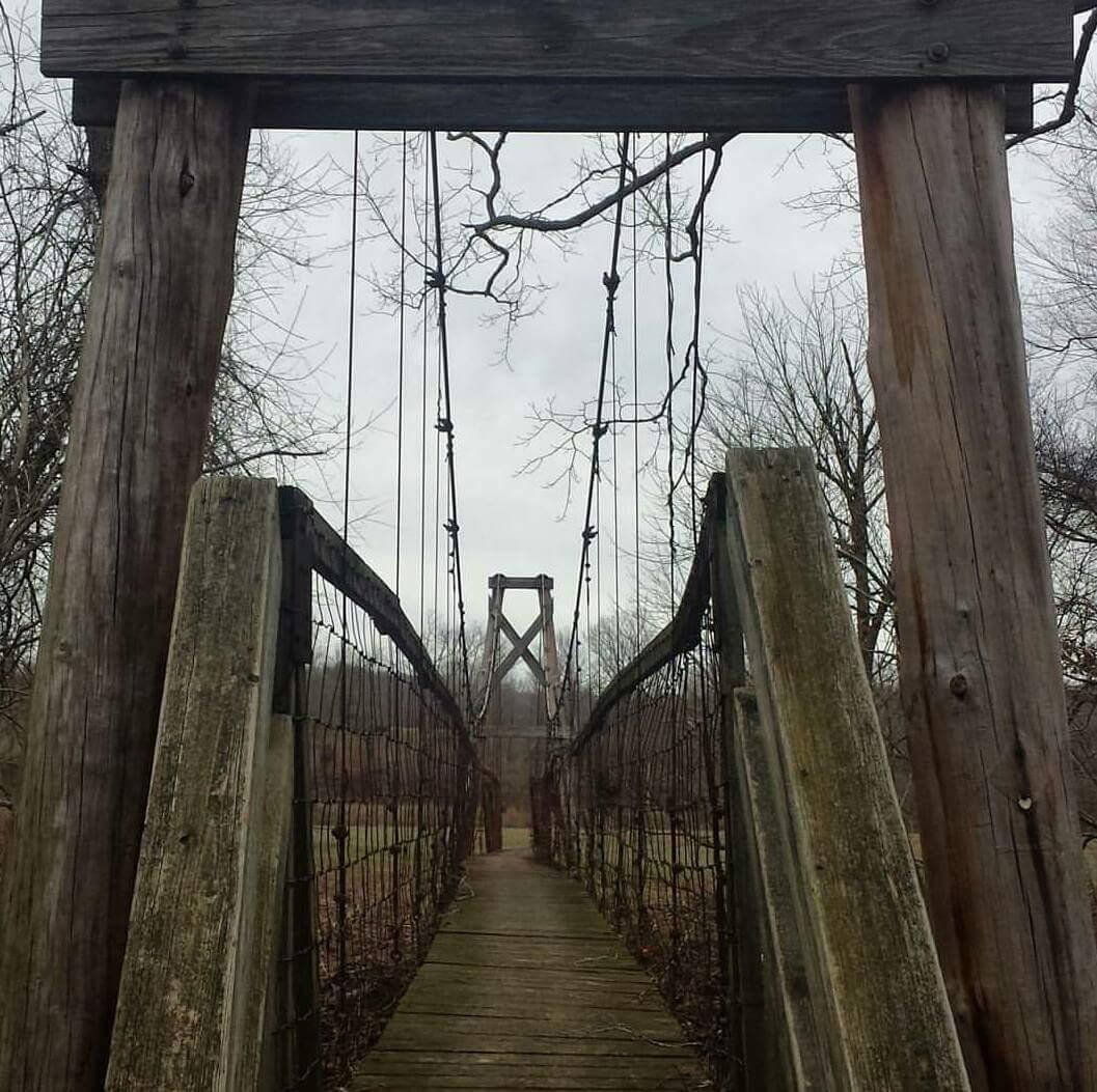 Suspension bridge made of wood and cables, leading into a wooded area with bare trees in the background under a cloudy sky. Cave-run Lake mountain bike trail.