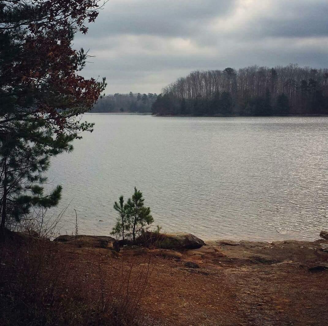 A tranquil lake scene under a cloudy sky, featuring calm waters in the foreground with rocky shores and sparse vegetation. In the background, trees line the opposite shore, creating a peaceful natural setting. Cave-run Lake mountain bike trail.