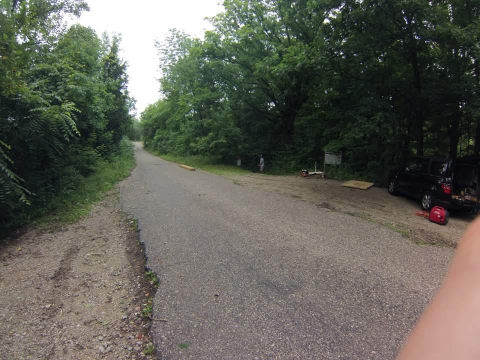 A partially paved road leading through a green, wooded area, with trees on either side. On the right, there is a small parking area with a black SUV and some outdoor equipment. A person is visible in the background, near the edge of the woods. The sky is overcast, suggesting a cloudy day. Caesar Creek mountain bike trail.