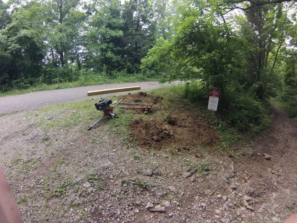 A partially dug area on a gravel path surrounded by trees, featuring a digging tool and a wooden beam nearby. There is a sign posted in the background on the right side, indicating the area. Caesar Creek mountain bike trail.