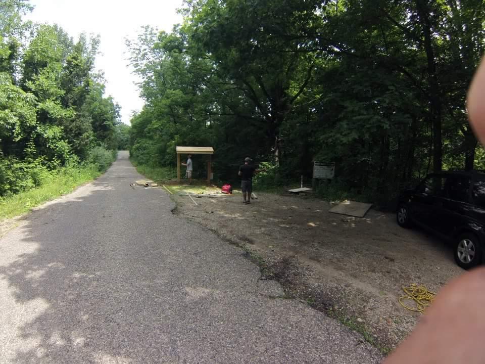 A partially paved road lined with greenery leads into a wooded area. In the foreground, two individuals are working near a newly constructed wooden structure that appears to be a shelter or signpost. A parked black car is visible on the gravel area to the right, with tools scattered nearby. Bright sunlight filters through the trees, creating a natural setting. Caesar Creek mountain bike trail.