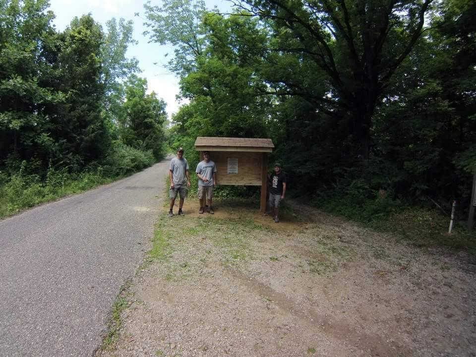 Three individuals stand near a wooden information kiosk beside a gravel road surrounded by lush green trees. The setting is a natural area, indicating a recreational or hiking spot. The sky is partly cloudy, suggesting a pleasant day. Caesar Creek mountain bike trail.