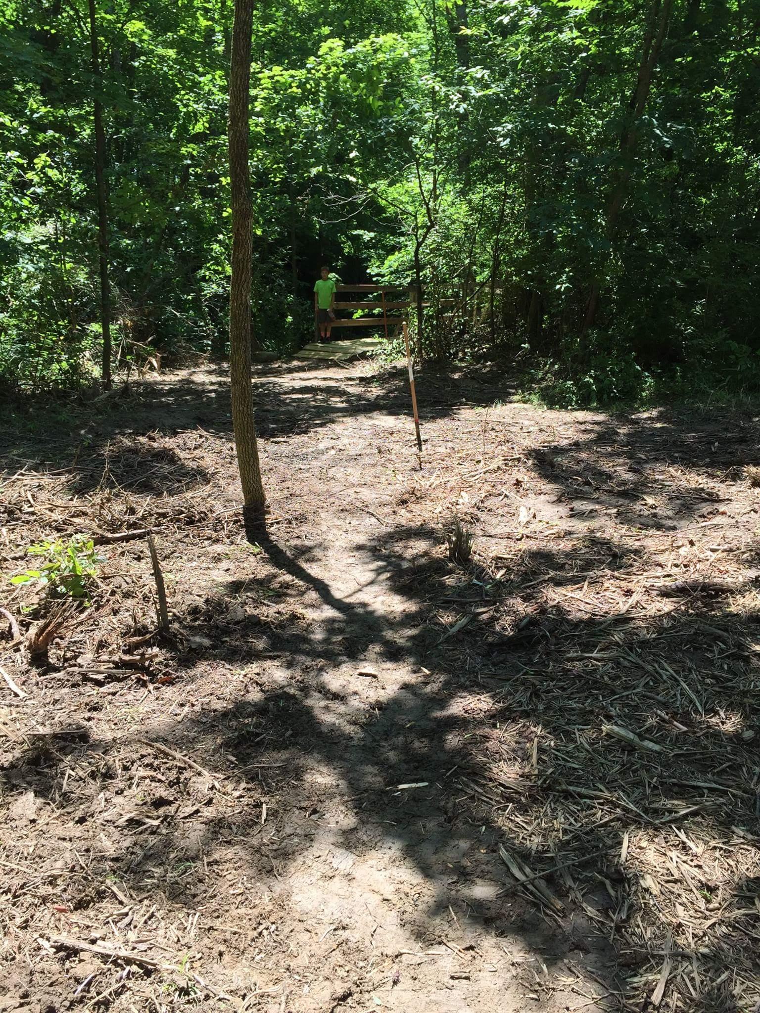 A sunlit path leading through a wooded area, with a clearing in the foreground and dense greenery surrounding it. In the background, a child wearing a green shirt stands near a wooden bridge. The ground is mostly bare with some scattered brush and small plants. Caesar Creek mountain bike trail.