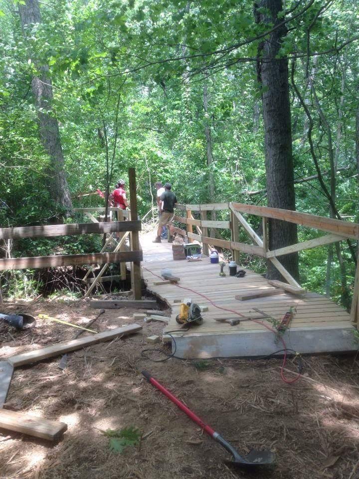 Construction of a wooden bridge in a forested area, with two people working on it. The scene includes wooden planks, tools on the ground, and greenery surrounding the area. Caesar Creek mountain bike trail.