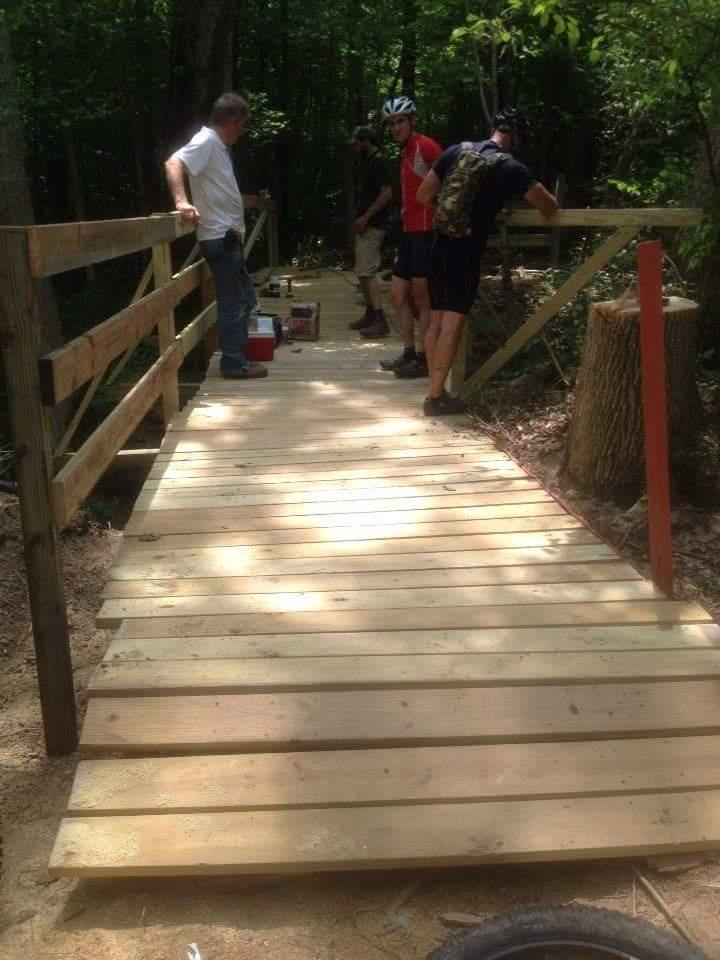 A group of individuals working together on the construction of a wooden bridge in a forested area. The bridge, made of wooden planks, is partially finished and surrounded by trees, with tools and materials visible nearby. Two of the people are inspecting the structure, while others are engaged in building tasks. Caesar Creek mountain bike trail.