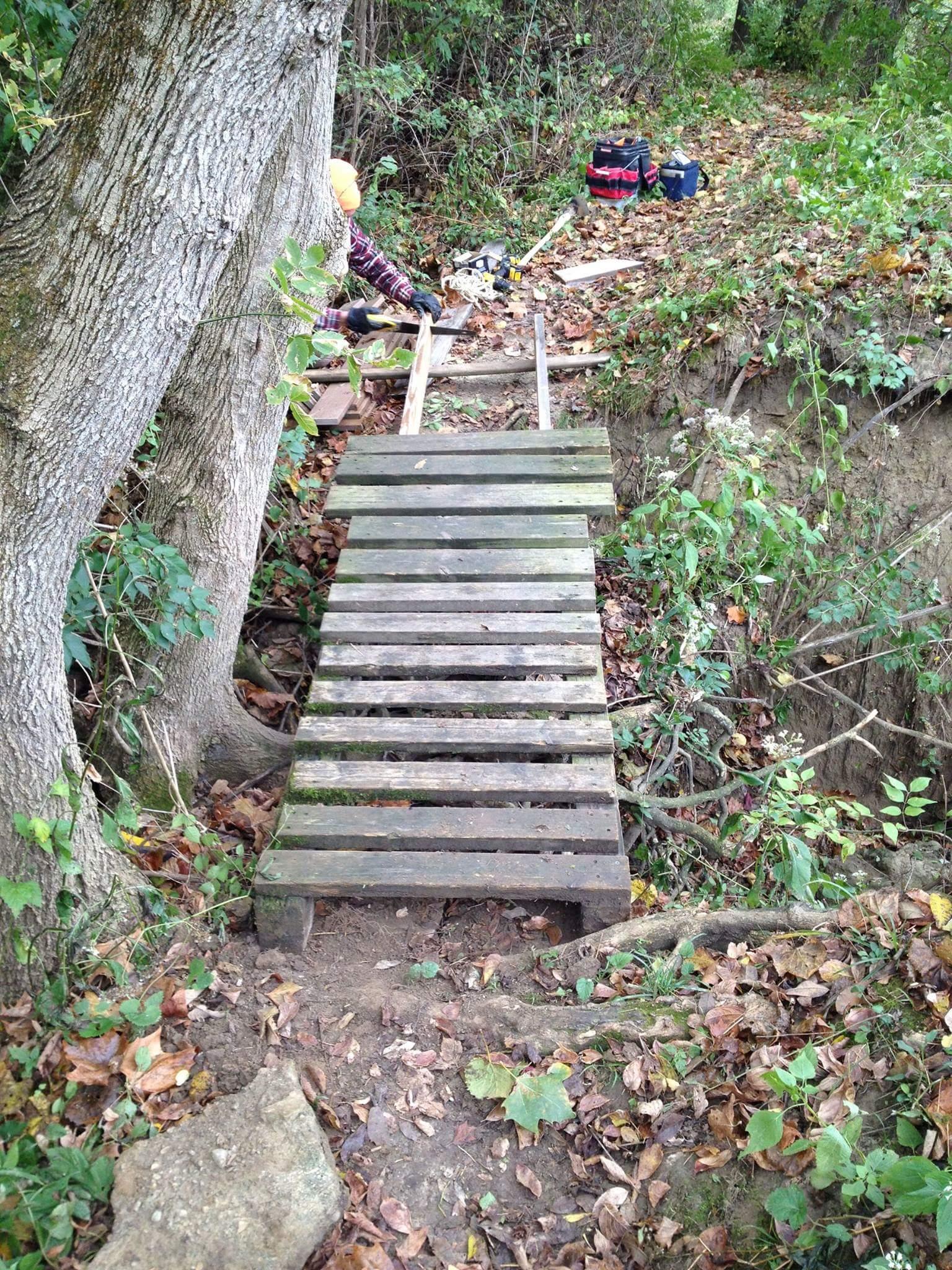 A wooden bridge sits over a small gap in a wooded area, surrounded by fallen leaves and plants. In the background, a person is working on a nearby structure, with tools and materials scattered around. The scene is quiet and natural, showcasing the effort involved in creating a path through the landscape. Caesar Creek mountain bike trail.