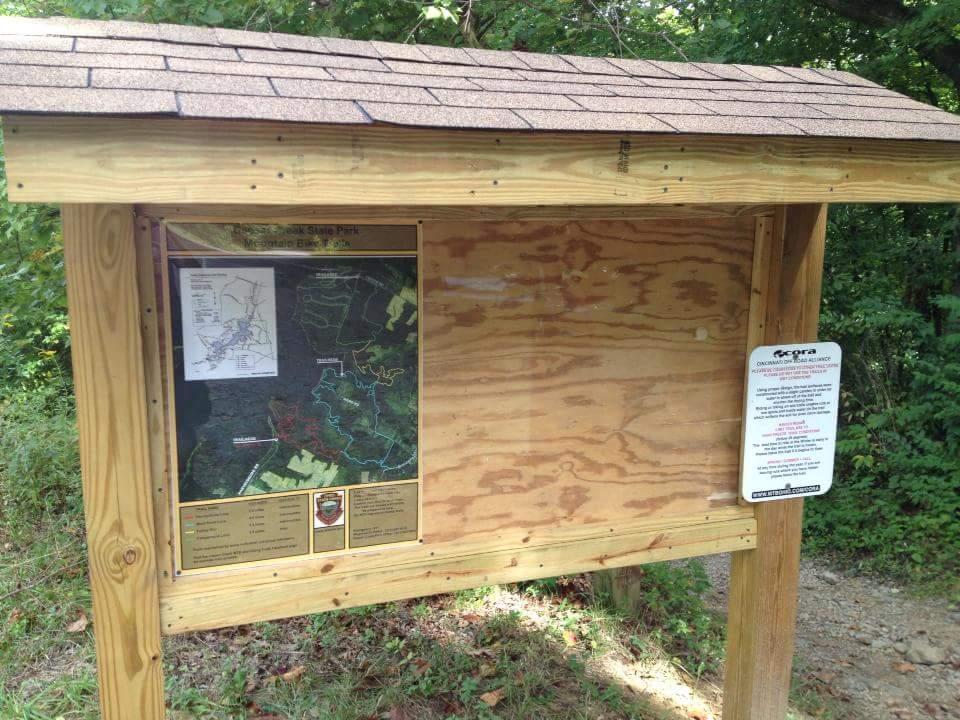 Wooden information kiosk at a trailhead featuring a map of the area, surrounded by greenery. The kiosk has a sloped roof and includes various informational postings about the park and surrounding trails. Caesar Creek mountain bike trail.