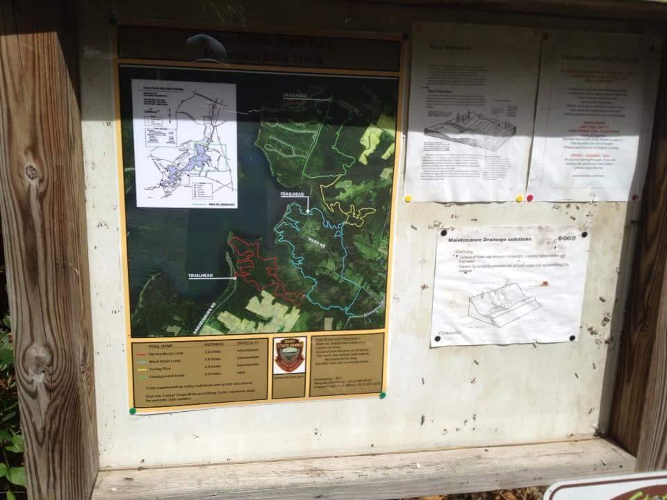 A wooden sign displaying a detailed map of the Hudson Highlands Bike Trail, including information about trail names, lengths, and difficulty levels. The map highlights various bike routes in a green landscape, with additional informational sheets about trail maintenance and safety posted nearby. Caesar Creek mountain bike trail.