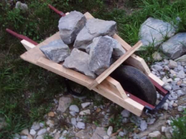 A wooden wheelbarrow filled with several large rocks, resting on a gravel surface surrounded by grass. The wheelbarrow features a single wheel and wooden handles, with a visible tire. Devou Park mountain bike trail.