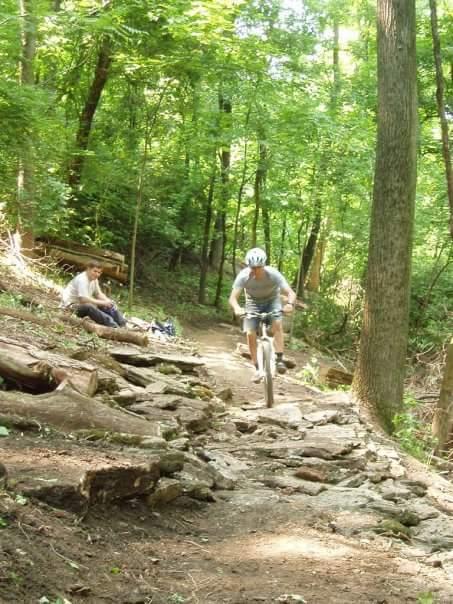 A cyclist navigates a rocky trail in a lush green forest, while a person sits on the side observing. The scene captures the excitement of mountain biking amidst nature, with sunlight filtering through the trees. Devou Park mountain bike trail.