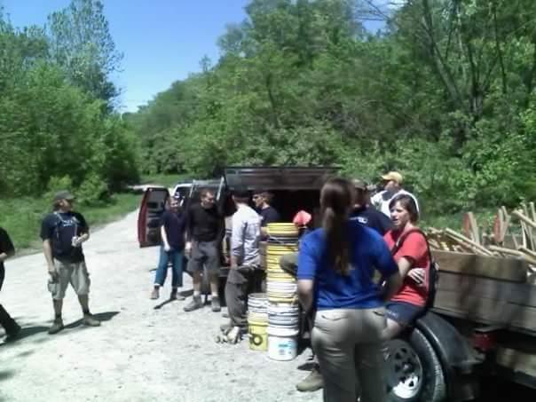 A group of people gathers by a pickup truck on a gravel road surrounded by lush greenery. They are engaged in conversation, with some individuals standing near large buckets and tools in the truck bed. The scene takes place on a clear, sunny day, reflecting a collaborative outdoor environment. Devou Park mountain bike trail.