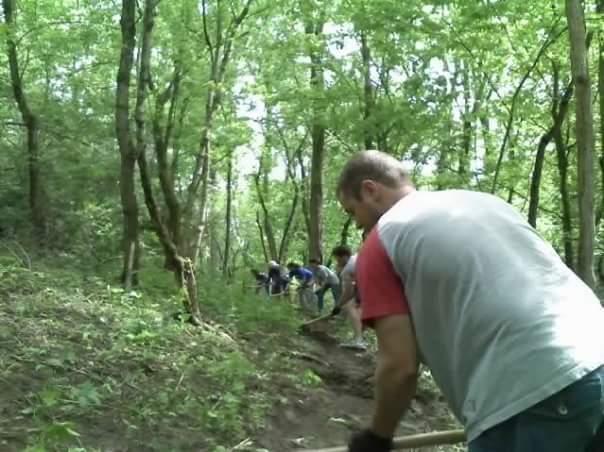 A group of people working together in a wooded area, engaged in outdoor maintenance activities. In the foreground, a man leans forward, using a tool to dig or move earth, while several individuals can be seen behind him, participating in the same task among the trees and greenery. Devou Park mountain bike trail.