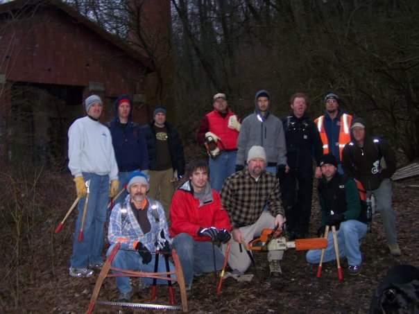 A group of twelve men posed outdoors in a wooded area, with an old building in the background. They are dressed in warm clothing suitable for outdoor work and are holding various tools, including a chainsaw, saw, and hand tools. The scene appears to be a gathering for a communal project or event. Devou Park mountain bike trail.