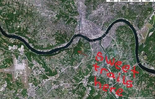 Aerial view of a city and surrounding landscape, featuring a winding river and various trails. The phrase "Sweet trails here" is prominently written in red text across the image. An airport is visible in the lower part of the image. Devou Park mountain bike trail.