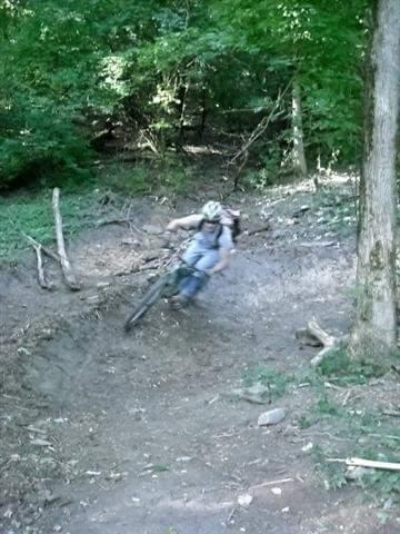 A person riding a mountain bike on a dirt trail through a wooded area, leaning into a turn surrounded by trees and foliage. Devou Park mountain bike trail.