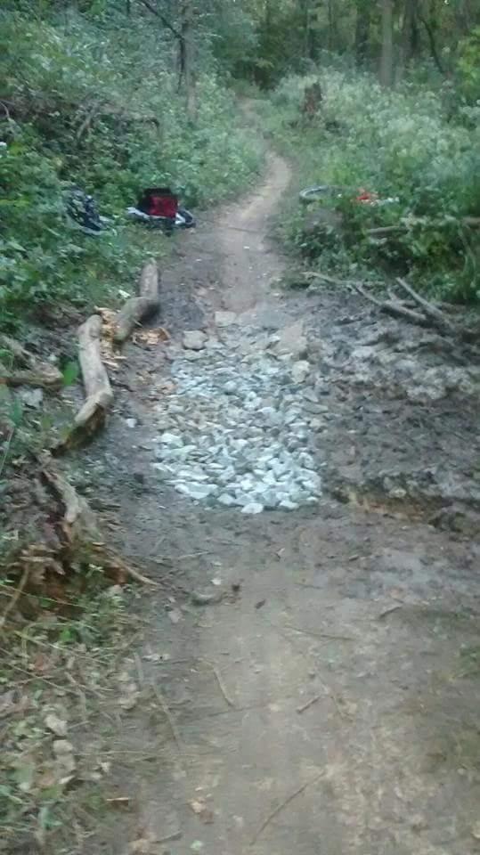 A narrow dirt path in a wooded area, featuring a section of gravel laid to improve drainage or footing. Surrounding greenery includes trees and shrubs, with a few personal items visible in the background. The trail appears slightly muddy and uneven. Devou Park mountain bike trail.