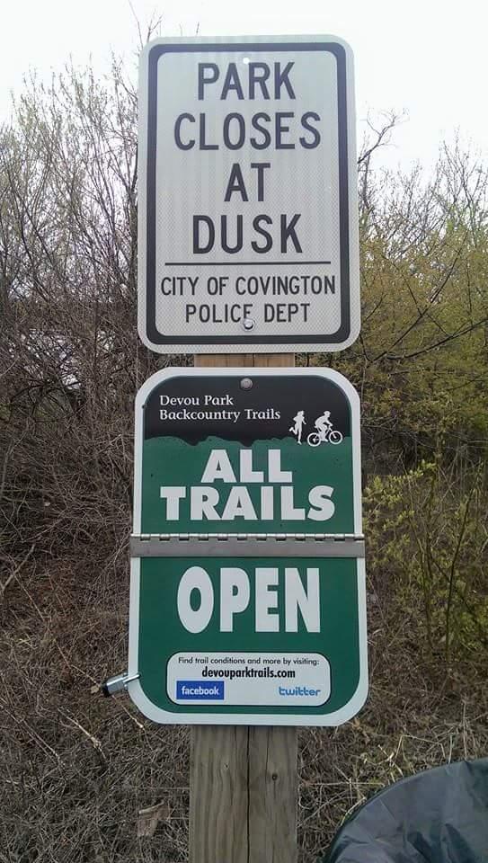 Signage indicating park regulations and trail information. The top sign states "PARK CLOSES AT DUSK," issued by the City of Covington Police Department. The bottom sign, related to Devou Park Backcountry Trails, declares "ALL TRAILS OPEN," including a suggestion to find more information online. Devou Park mountain bike trail.