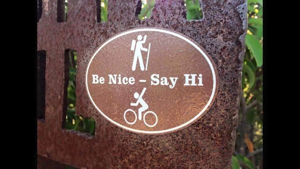 A brown sign featuring two iconographic figures: one representing a hiker and the other a cyclist. The text reads, "Be Nice - Say Hi." The sign is mounted on a rustic, textured background, emphasizing friendliness among outdoor enthusiasts. Devou Park mountain bike trail.