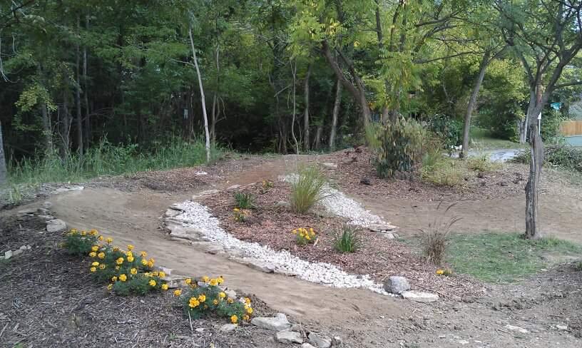 A landscaped area featuring curved pathways bordered by small stones and mulch, surrounded by green trees. Bright yellow marigolds and various plants are planted along the pathways, adding color to the natural setting. Devou Park mountain bike trail.