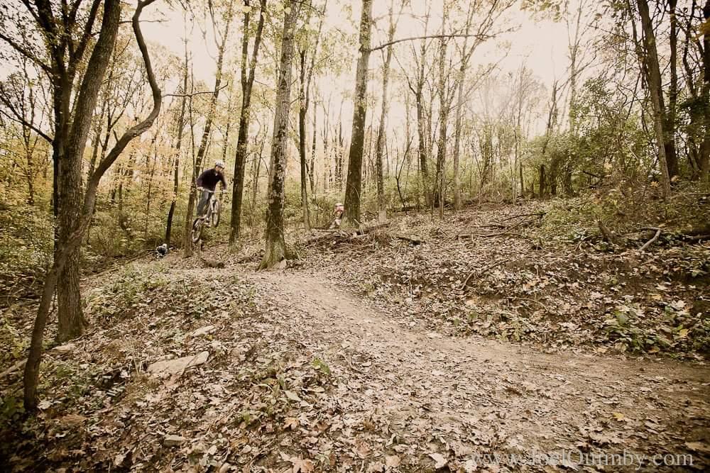 A mountain biker performing a jump on a dirt trail surrounded by trees, with autumn foliage in the background. The scene captures the excitement of outdoor biking in a forested area. Devou Park mountain bike trail.
