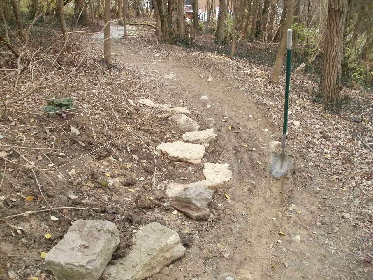 A dirt path in a wooded area, featuring a newly laid stone border. A shovel is stuck in the ground beside the path, and there are dry leaves and twigs scattered around. Trees and greenery are visible in the background. Devou Park mountain bike trail.