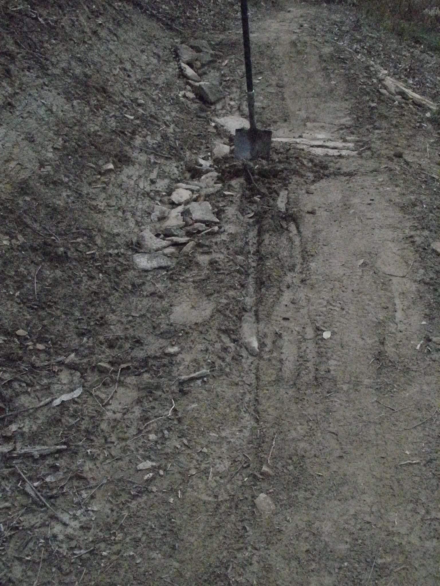 A dirt path running alongside a steep embankment, with scattered rocks and a shovel standing upright in the ground. The area appears to be recently disturbed, with visible tire tracks and loose soil, indicating recent work or maintenance. Devou Park mountain bike trail.