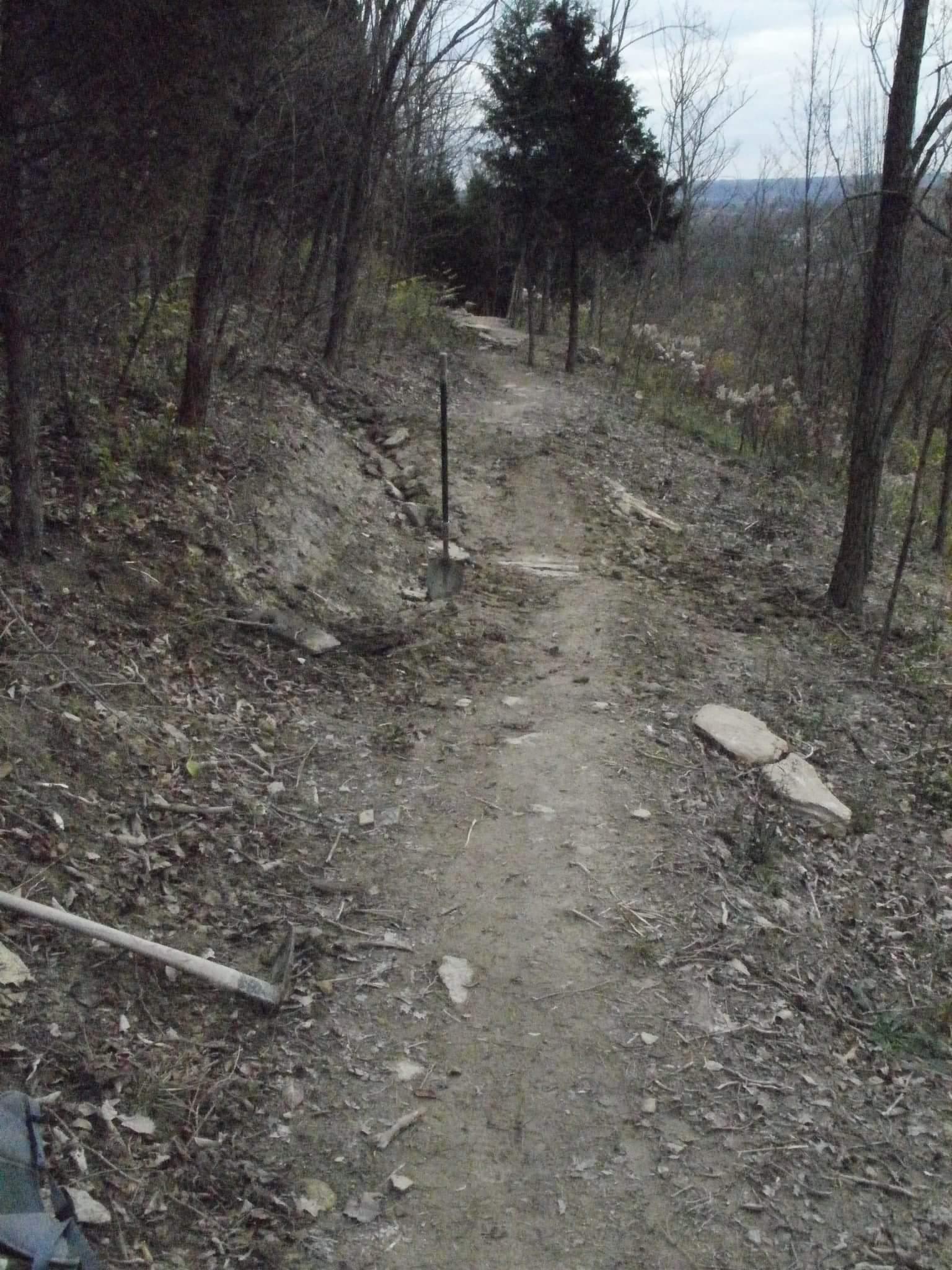 A narrow dirt path winding through a wooded area, with clearing vegetation and scattered rocks along the sides. A shovel and a hoe are positioned on the ground, indicating recent landscaping or trail work. Bare trees with few leaves are visible, suggesting it may be late fall or early spring. Devou Park mountain bike trail.