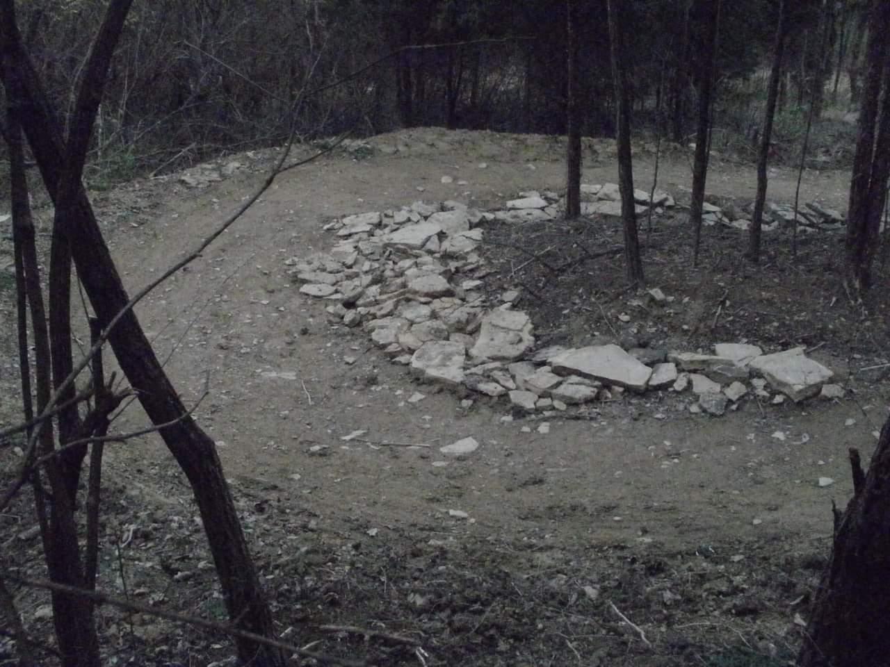 A winding dirt path in a wooded area, bordered by trees. The path features a circular arrangement of large stones along one side, indicating a potential resting spot or trail marker. The lighting suggests either early morning or late afternoon, creating a serene and natural atmosphere. Devou Park mountain bike trail.