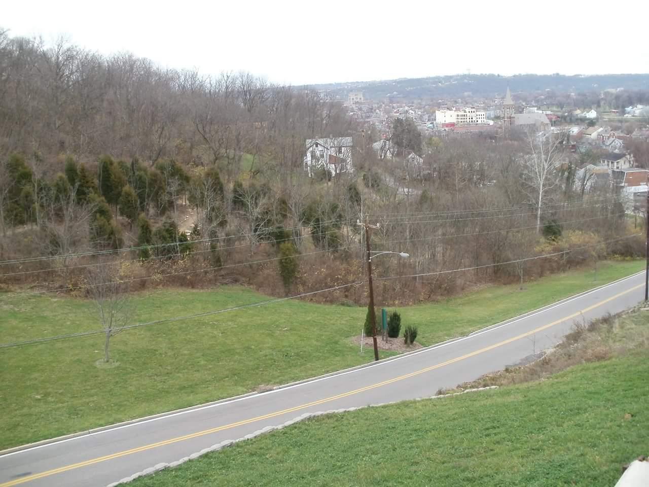 A scenic view from a hillside overlooking a rural area with trees and houses, featuring a winding road in the foreground. The landscape is partially bare, indicating early spring or late autumn, and shows a mix of greenery and utility poles. In the distance, a small town with buildings is visible against a cloudy sky. Devou Park mountain bike trail.