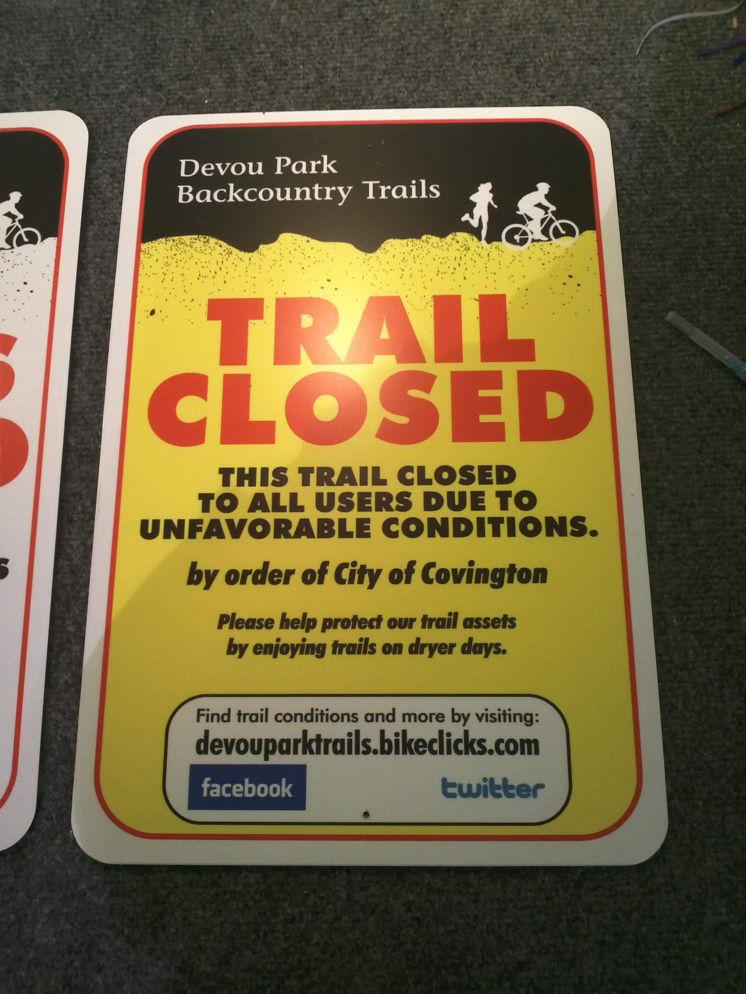 A vibrant trail closure sign for Devou Park Backcountry Trails, featuring a bold "TRAIL CLOSED" message in red letters. The sign indicates that the trail is closed to all users due to unfavorable conditions, as ordered by the City of Covington. Additional information encourages users to help protect the trails by visiting during dryer days and provides a website for more trail conditions, along with social media icons for Facebook and Twitter. Devou Park mountain bike trail.