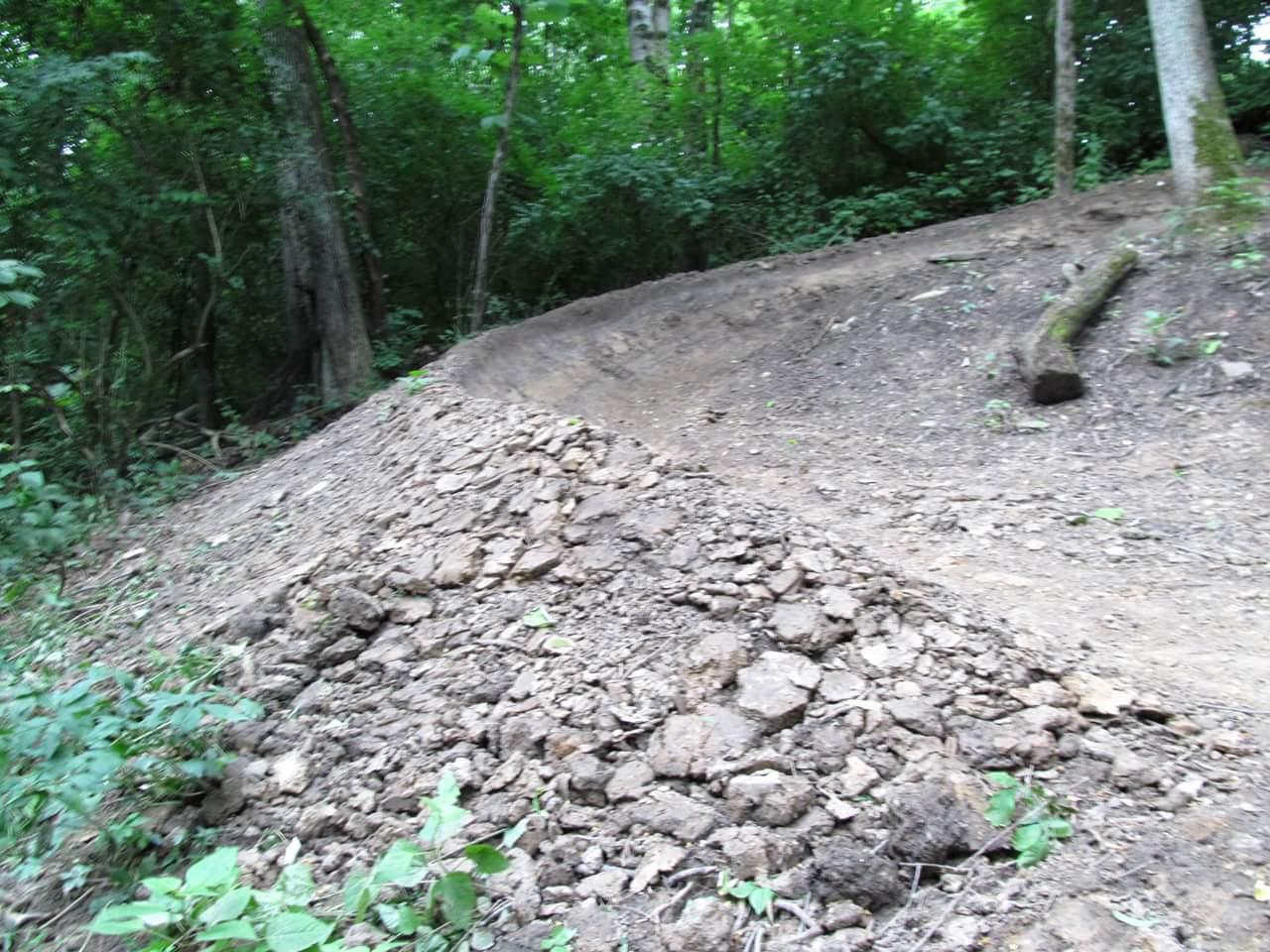 A dirt path winding through a wooded area, featuring a raised mound of soil on the left side, indicating recent trail work or landscaping. Lush greenery and trees surround the path, creating a natural outdoor setting. Devou Park mountain bike trail.