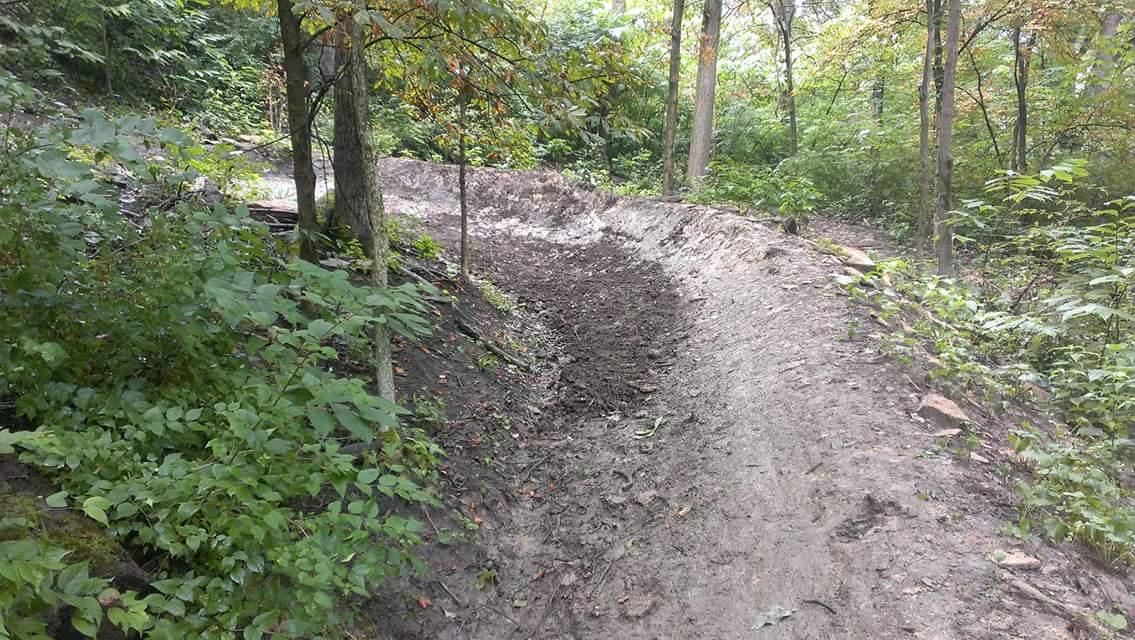 A winding dirt path through a wooded area, surrounded by trees and lush green vegetation. The trail is slightly muddy and curves to the right, with patches of light filtering through the leaves above. Devou Park mountain bike trail.