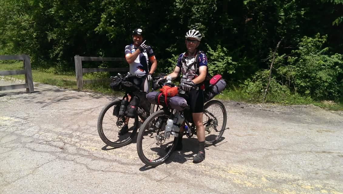Two cyclists pose beside their bicycles on a sunny day, surrounded by greenery. Each bike is loaded with gear, indicating they are on an outdoor adventure. One cyclist gives a thumbs-up, while the other stands with a relaxed expression, both wearing cycling jerseys and helmets. Devou Park mountain bike trail.