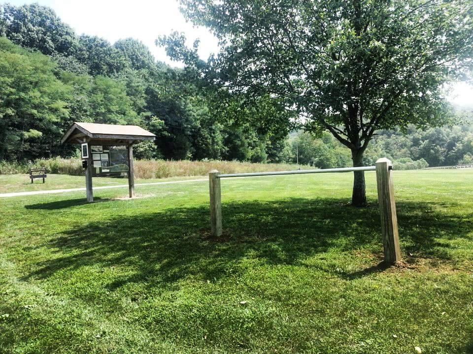 A grassy park featuring a wooden information kiosk with a roof, adjacent to a bench and a tree. The scene is surrounded by lush greenery and a pathway, with a clear sky above. Versailles State Park mountain bike trail.