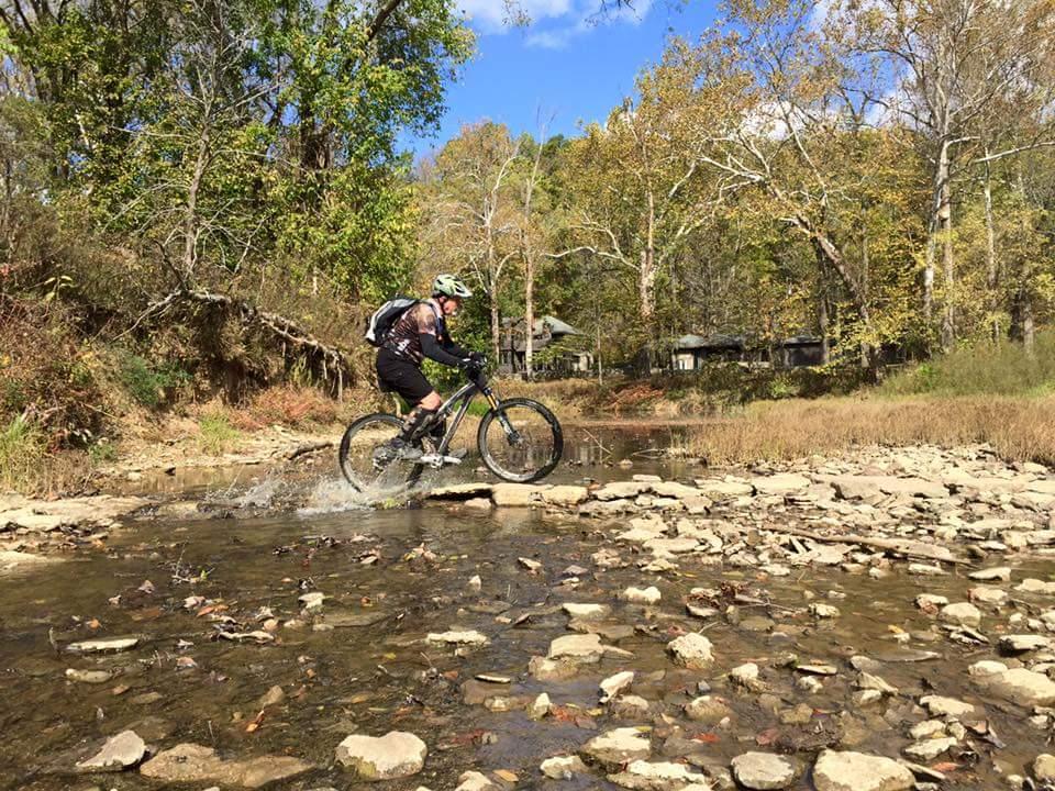 A mountain biker navigates across a rocky creek, splashing through the water. The scene is set in a wooded area with colorful autumn foliage and clear blue skies. Versailles State Park mountain bike trail.