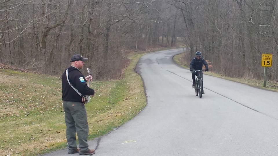 A man playing a banjo stands on a road while a cyclist rides by. The scene is set in a wooded area with bare trees, and a speed limit sign indicating 15 mph is visible nearby. Versailles State Park mountain bike trail.
