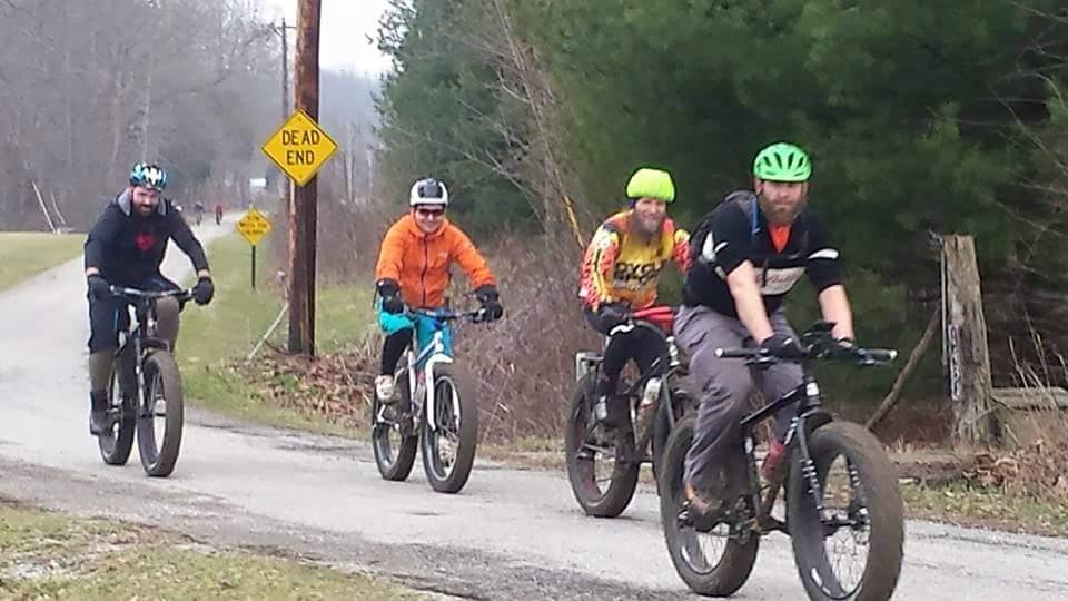 A group of four cyclists riding fat bikes along a rural road. The first cyclist is wearing a black hoodie and black pants, the second is in an orange jacket, the third wears a bright yellow helmet and colorful jersey, and the fourth has a green helmet and black clothing. A "Dead End" sign is visible in the background, along with trees lining the road. Versailles State Park mountain bike trail.